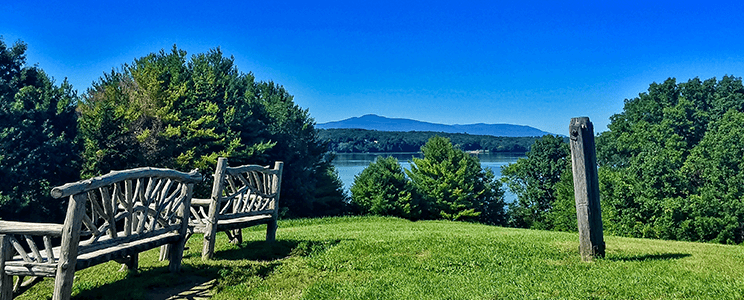 Photograph taken from a hill in the Hudson Valley in NYS with two benches, blue mountains, trees, & water in the background.